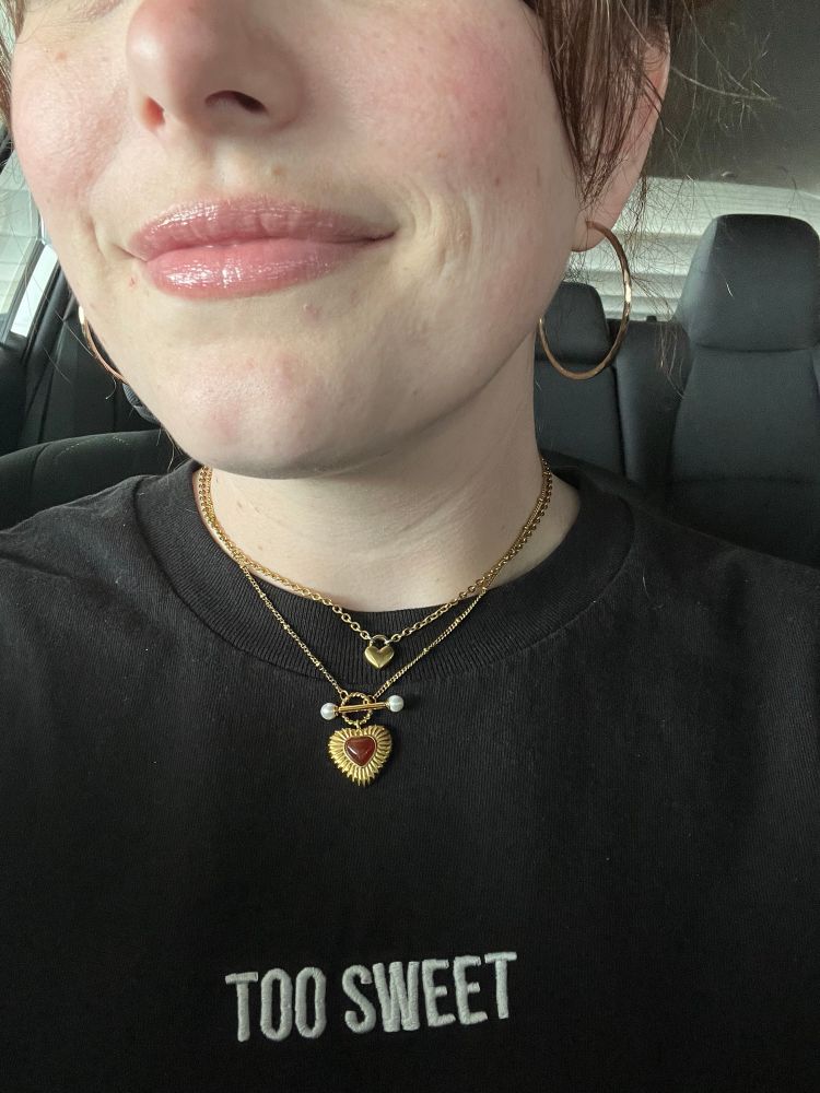A close up photo of a white woman from the nose down. She wears a black “Too Sweet” shirt and 2 gold heart necklaces. 