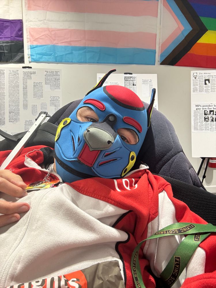 Disabled pup in a blue and red hood, in front of non-binary, trans, and Progress flags on a white wall. There are also three placards with newspaper clippings highlighting attitudes about LGBTQ+ people from years past