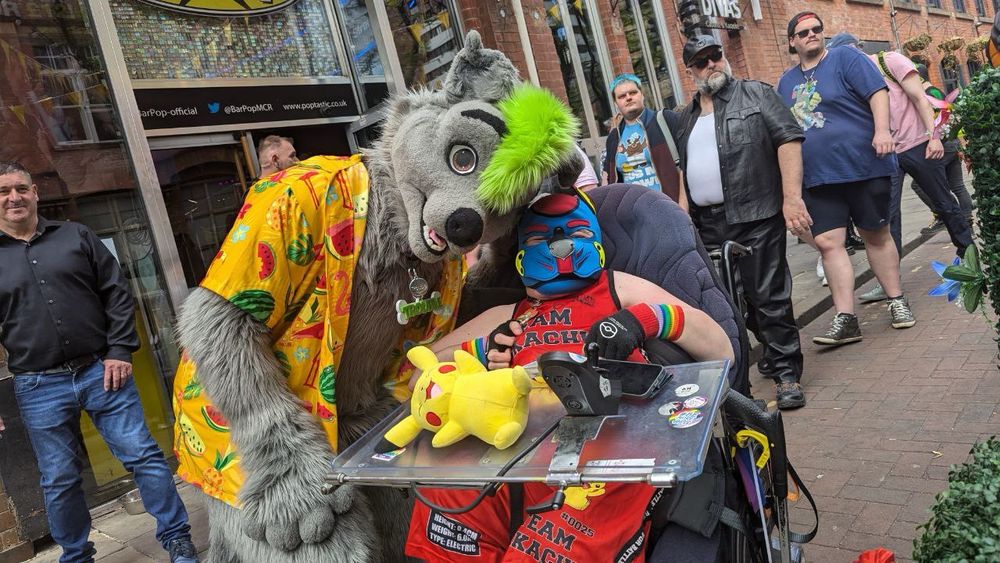 Disabled pup in a blue and red hood, wearing a red Pikachu sports outfit, with a grey and green furry in an orange Hawaiian shirt 