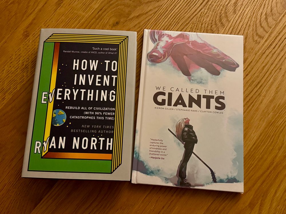 The books “How To Invent Everything” by Ryan North, and “We Called Them Giants” by Kieron Gillen, Stephanie Hans, and Clayton Cowles sitting on a table, shot from above. 