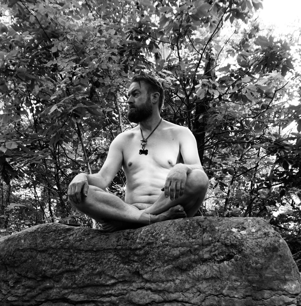 A monochromatic photo of Me nude and sitting cross legged on a rock ledge.