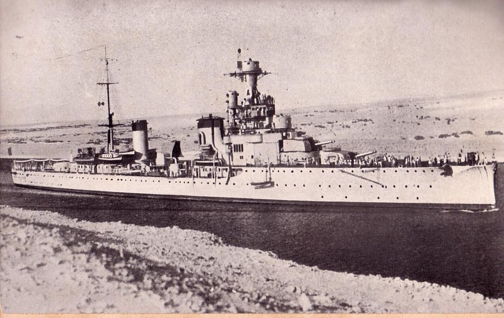 Italian light cruiser Bartolomeo Colleoni transiting the Suez Canal on return from the China station. The picture is black and white, taken at an angle from the port bow. It shows the cruiser in the narrow body of water with the desert behind it.
