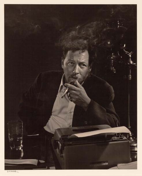 Playwright Tennessee Williams at his desk, leaning over a typewriter with a page in process, a glass to his right, smoking, looking up and at an angle from the camera.
