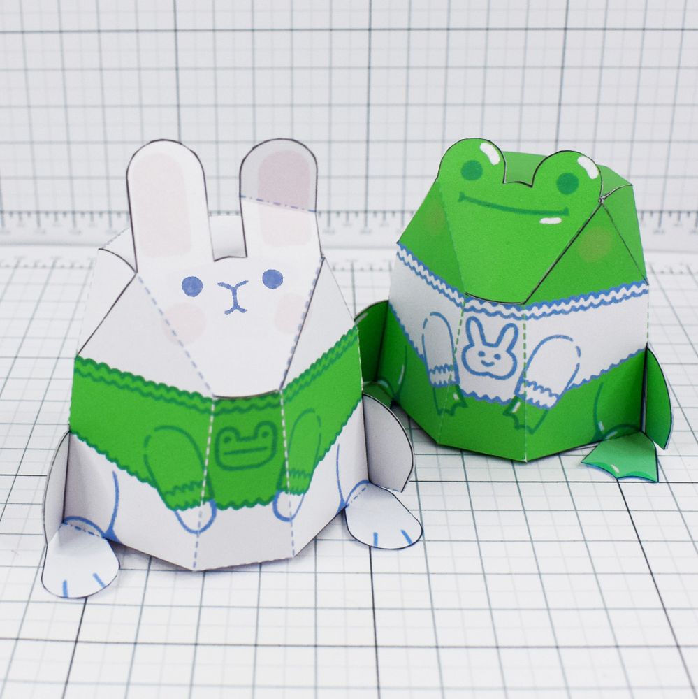 A photo of two papercrafts, one of a rabbit wearing a jumper with a frog on it, one of a frog wearing a jumper with a rabbit on it.