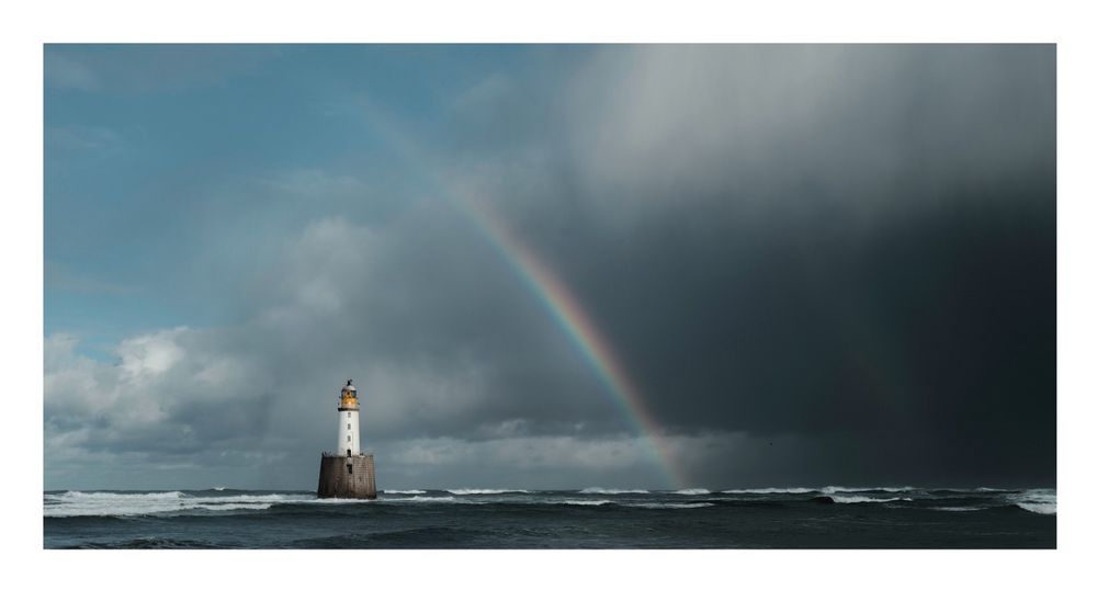 Image of a lighthouse in the ocean with dark clouds and a vibrant rainbow.