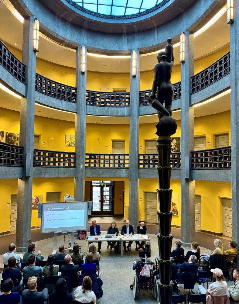 Panoramafoto eines sehr hohen Saales, mit einer ebenfalls sehr hohen Skulptur, unten das Publikum in Reihen und an einem Podium vier Personen.