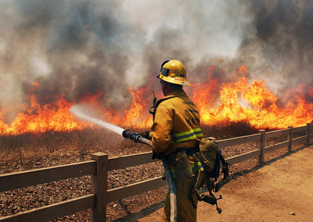 A fireman in emergency uniform, using a firehose to extinguish a roaring flame that is burning up a dry field. Credit: LAFD Foundation