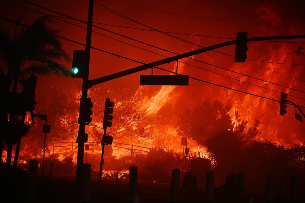 A wall of flame engulfs an intersection, darkening the sky. Against the red backdrop, the silhouette of traffic lights are visible, along with the green "go" light. Credit: Robyn Beck/AFP