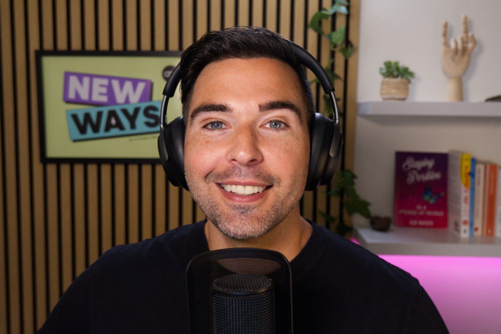 A person in a black t-shirt wearing headphones smiling as they speak into a microphone. Behind them is a sign that reads New Ways.