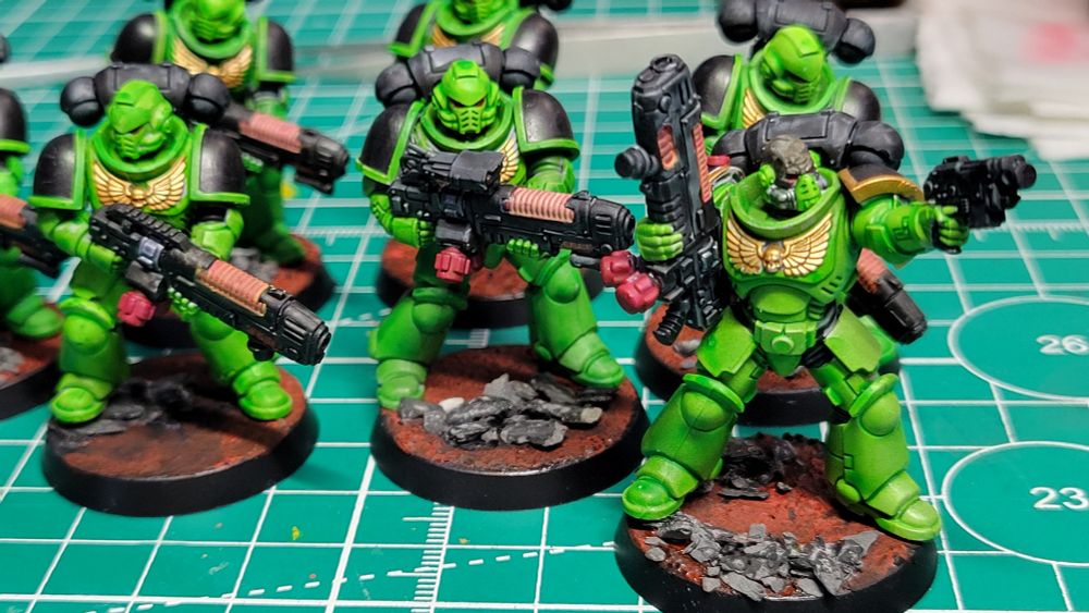 A close-up picture of 6 Warhammer Hellblaster minis painted as part of the Salamanders Space Marines chapter. They have green armor, black pauldrons, and are standing on red bases with grey rocks. One model is not wearing a helmet and pointing a Bolt Pistol with his left hand