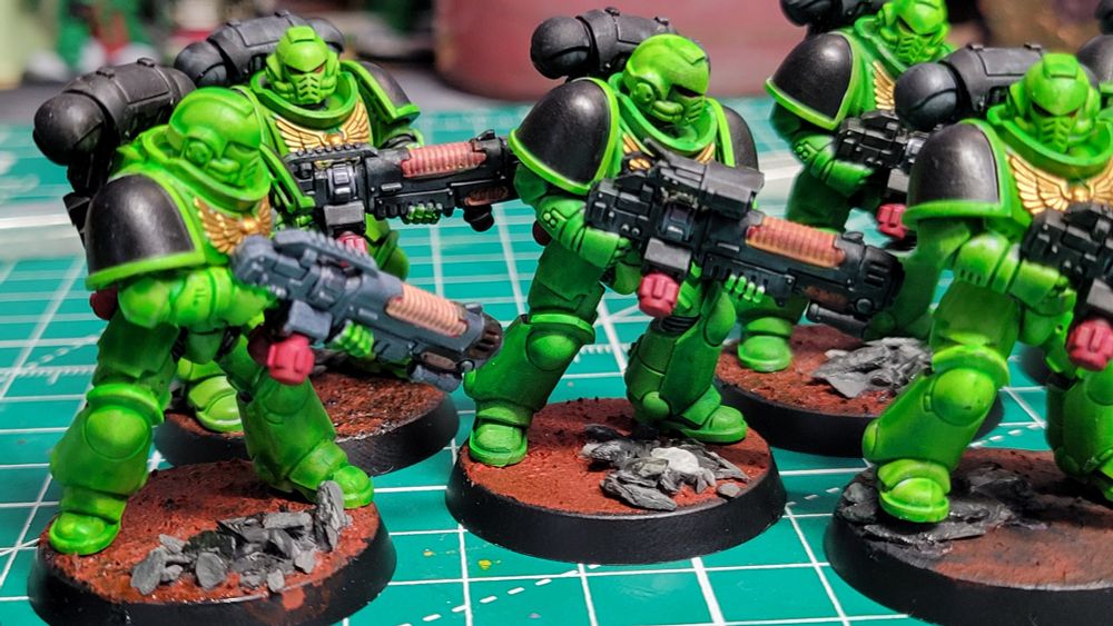 A close-up picture of 5 Warhammer Hellblaster minis painted as part of the Salamanders Space Marines chapter. They have green armor, black pauldrons, and are standing on red bases with grey rocks. Their Plasma Incinerators are black with bright red fuel tanks on the bottom and glowing orange plasma coils on top. The plasma coils are painted very badly.