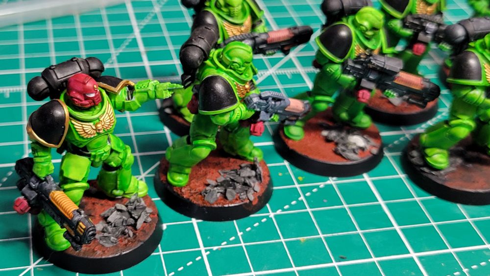 A picture of 5 Warhammer Hellblaster minis painted as part of the Salamanders Space Marines chapter. They have green armor, black pauldrons, and are standing on red bases with grey rocks. The Sergeant model has a red helmet and is pointing with his left hand.