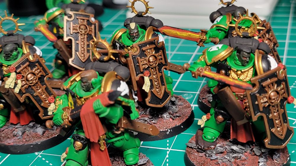 A close-up picture of six Bladeguard Veteran models. They are painted as part of the Salamanders chapter. They have green armor with gold trim. They are carrying power swords that are glowing red from the inside, fading to black at the edges. Their shields are black with bronze trim. They are all helmet-less and have dark gray skin. They are standing on bases with dusty red soil and large grey rocks.