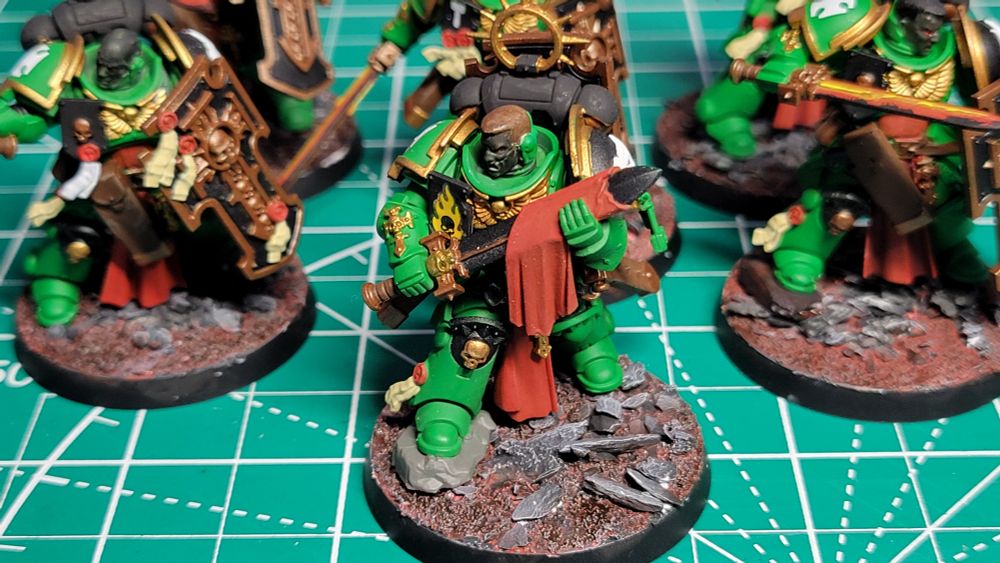A close-up picture of a Bladeguard Veteran Sargeant model. He is painted as part of the Salamanders chapter. He has green armor with gold trim. His power sword is held in both hands across his chest with a red cloth draped over it. He is standing on a base with dusty red soil and large grey rocks.
