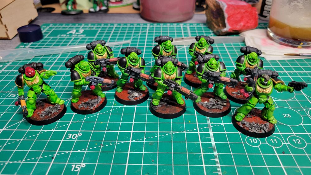 A picture of 10 Warhammer Hellblaster minis painted as part of the Salamanders Space Marines chapter. They have green armor, black pauldrons, and are standing on red bases with grey rocks. The Sergeant model has a red helmet.