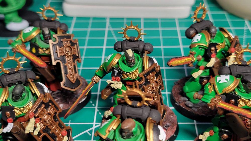 A close-up picture of six Bladeguard Veteran models. They are painted as part of the Salamanders chapter. They have green armor with gold trim. They are carrying power swords that are glowing red from the inside, fading to black at the edges. Their shields are black with bronze trim. They are all helmet-less and have dark gray skin. They are standing on bases with dusty red soil and large grey rocks.