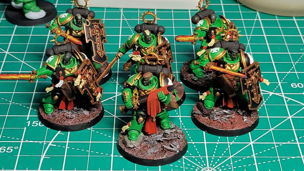 A picture of six Bladeguard Veteran models. They are painted as part of the Salamanders chapter. They have green armor with gold trim. They are carrying power swords that are glowing red from the inside, fading to black at the edges. Their shields are black with bronze trim. They are all helmet-less and have dark gray skin. They are standing on bases with dusty red soil and large grey rocks.