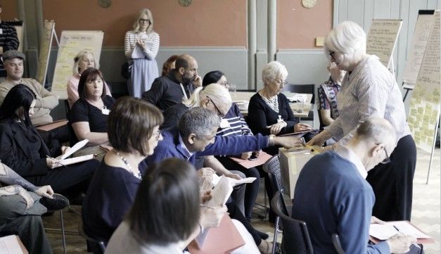 The Citizens Jury: A group of adults sit in a classroom-style setting, some reading papers while others interact. A woman at the front collects a vote from a seated man. Flipcharts with notes are visible in the background.