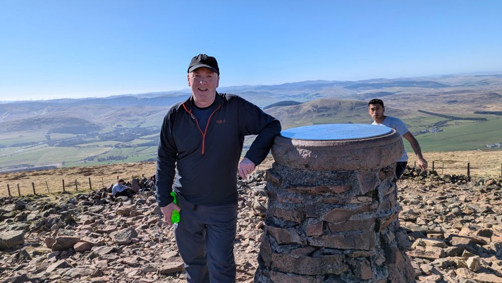 Man with a hat (me)leaning against the cairn at the top of Tinto Hill in South Lanarkshire. Rolling hills behind me. 