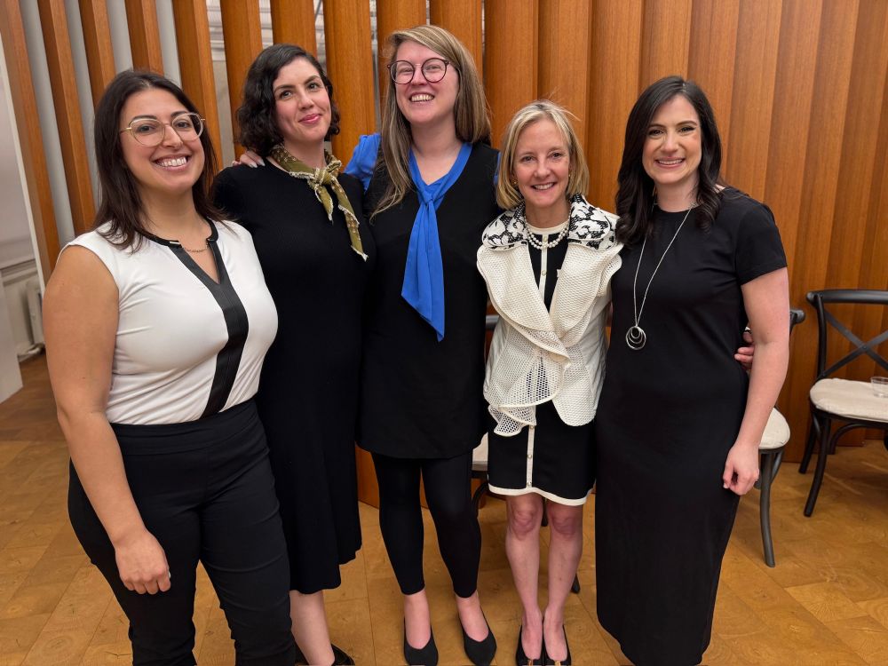 Five women standing in front of a brown paneled wall with their arms around each other smiling for the picture. 