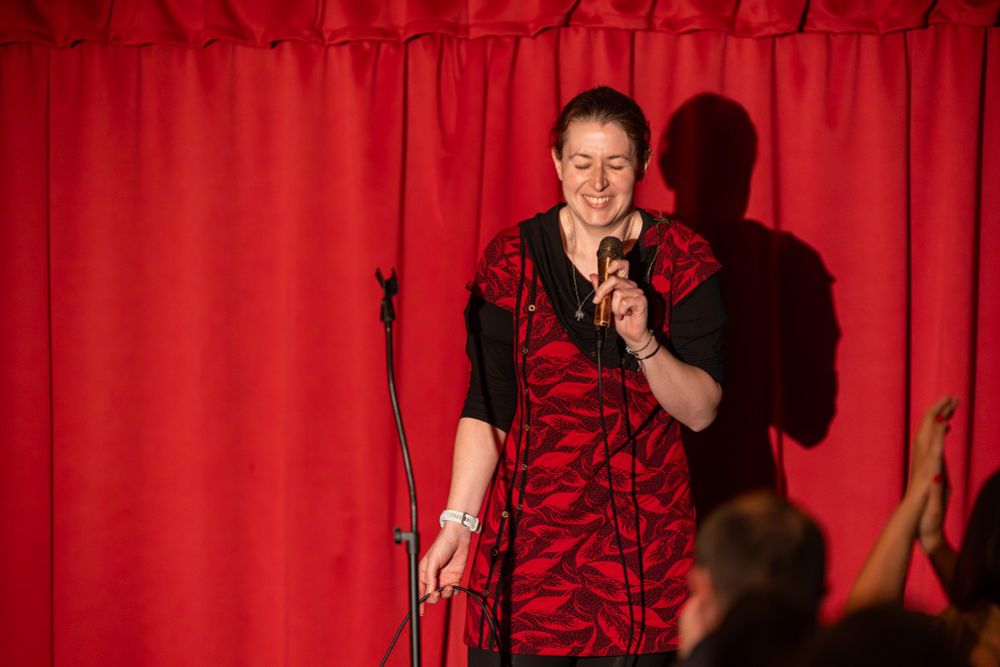 A laughing scientist pretending to be a comedian on stage dressed in a red tunic that is almost identical to the curtain behind. 
