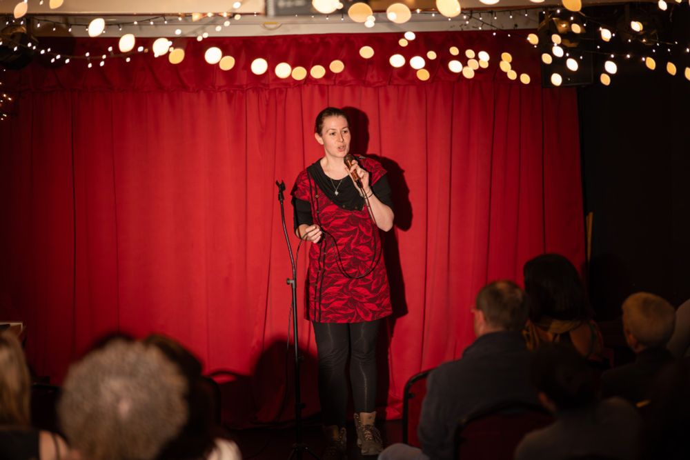 A scientist pretending to be a comedian on stage dressed in a red tunic that is almost identical to the curtain behind. 
