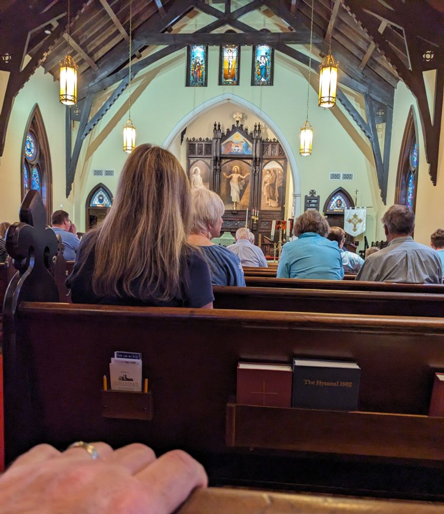 Picture of hand on pew from the back of an Episcopal church, facing images of Christ at the front of the church. 