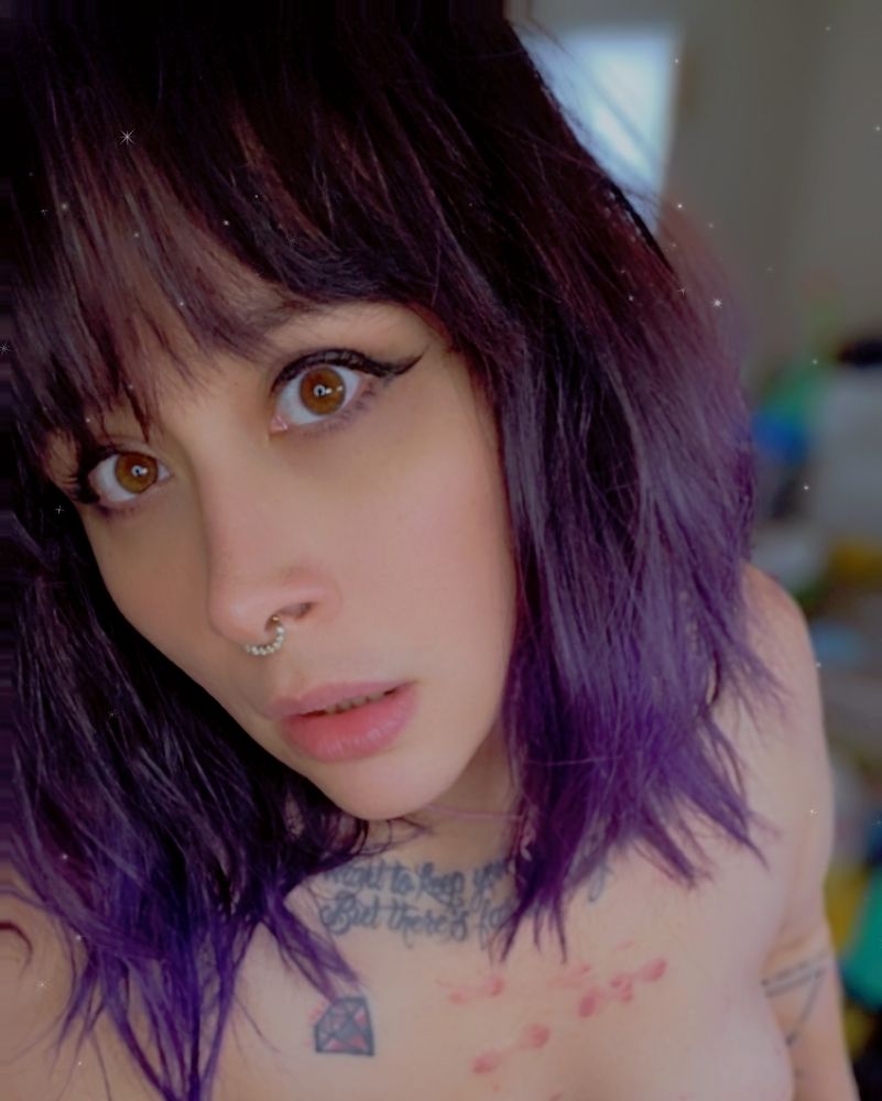 Messy wavy purple and black haired woman with chest tattoos staring intensely into the camera with big brown eyes