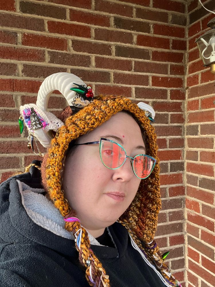 A selfie of a person with glasses, wearing a brown crochet hat with plastic horns that have been accessorized with beads and jewelry. 