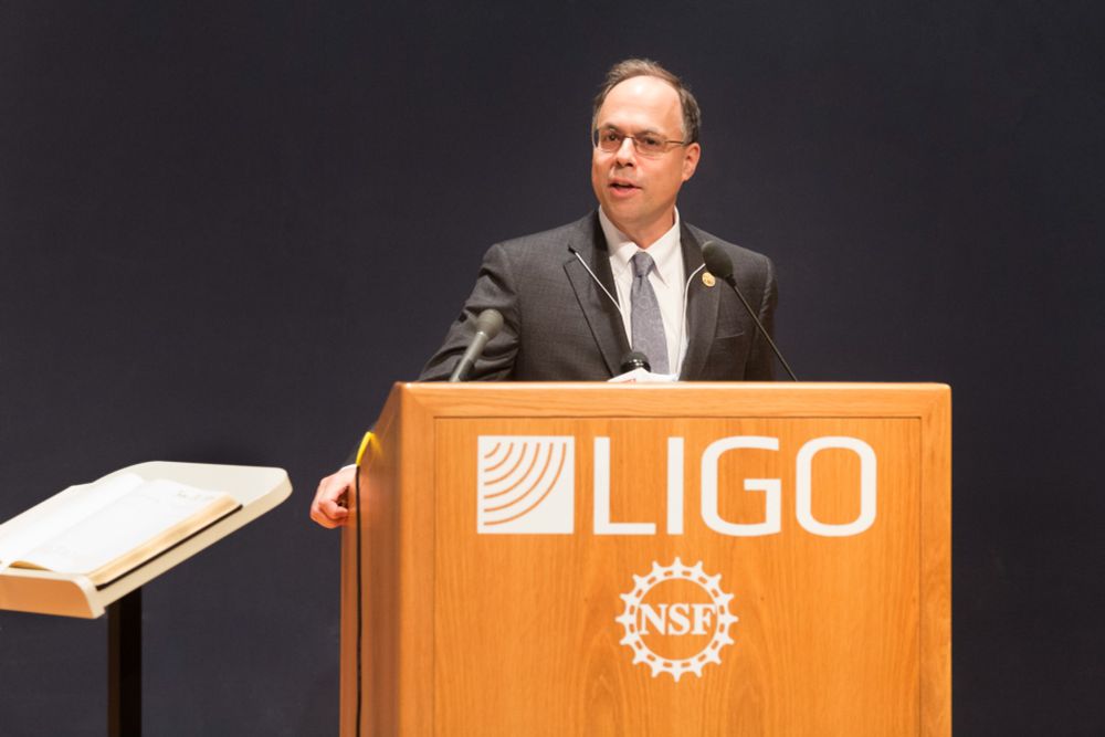 A photo of LSU professor and head of LIGO Joseph Giamie speaking at an event behind a wood lectern with LIGO and the National Science Foundations logos on the front.