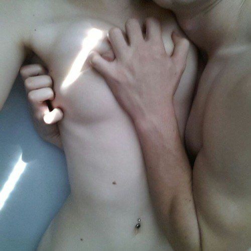 A man and woman embrace naked in bed, the man clutching the woman close to him, his left hand pressing her breasts to her chest. A shaft of sun light, cuts diagonally across her right breast. Subdued, barely coloured tones.
