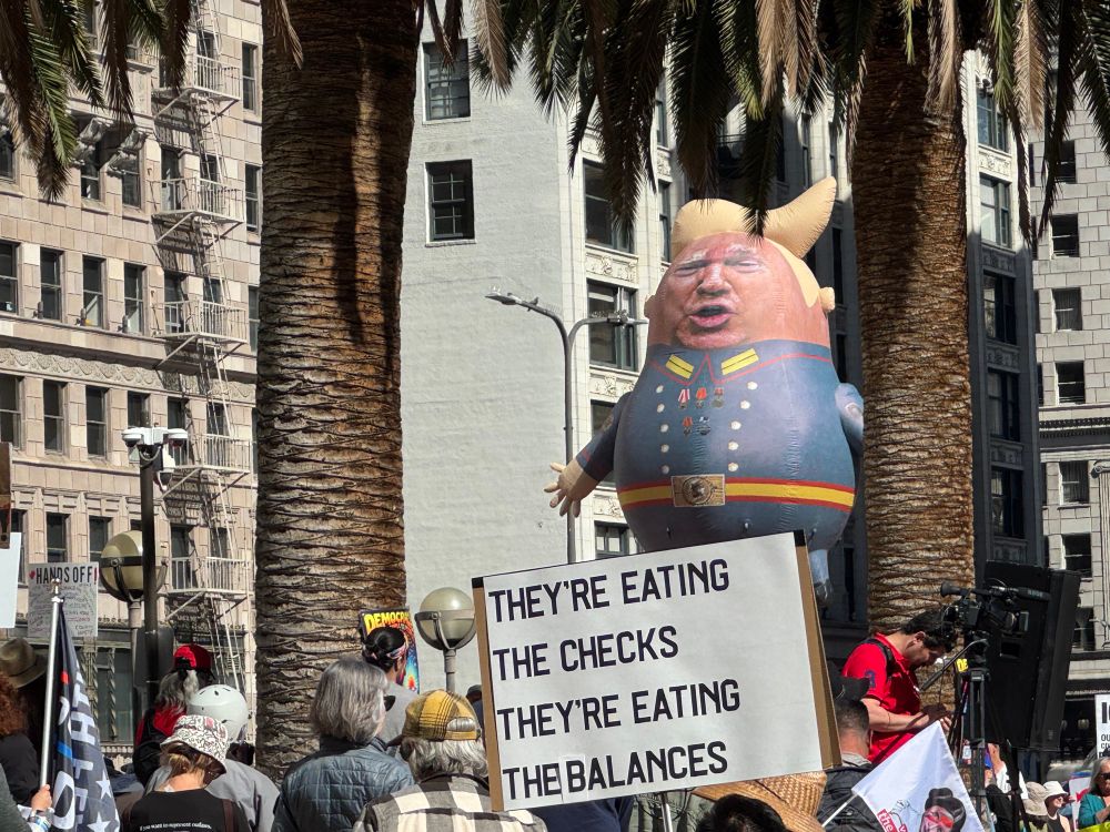 A giant dolly Polly trump balloon behind a sign reading “they’re eating the checks. They’re eating the balances”