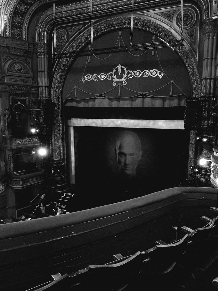 A grainy black and white photo of the stage in leeds grand theatre. The curtain is down with a photo of derren browns face on it
