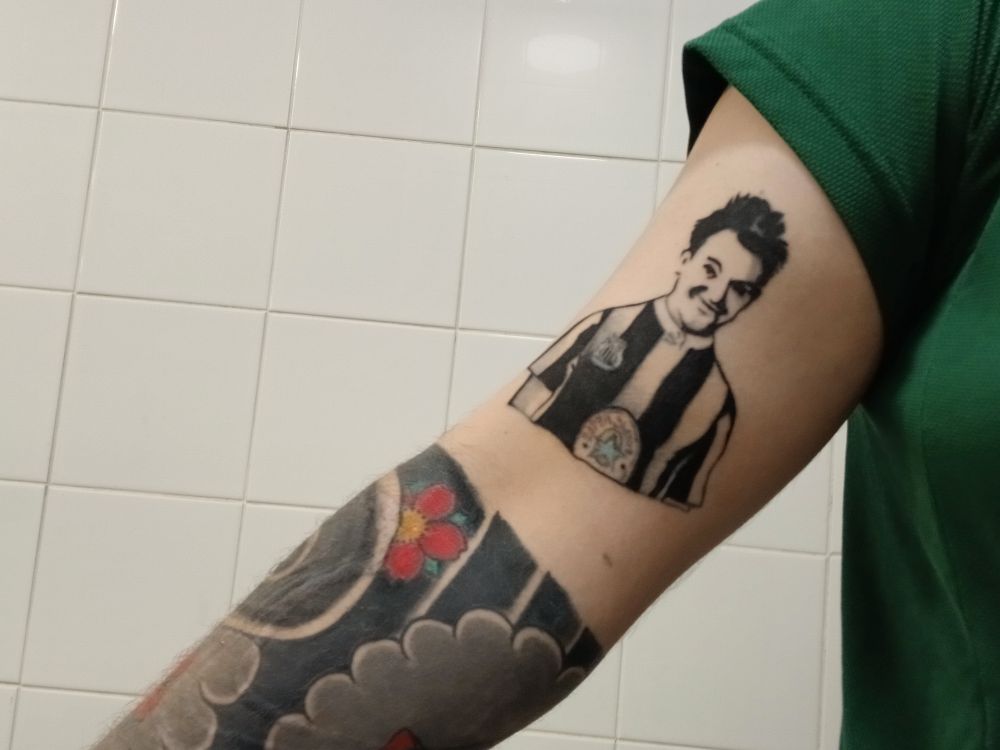 The same white arm, tattoos, and green t shirt, but in front of a white tiled wall