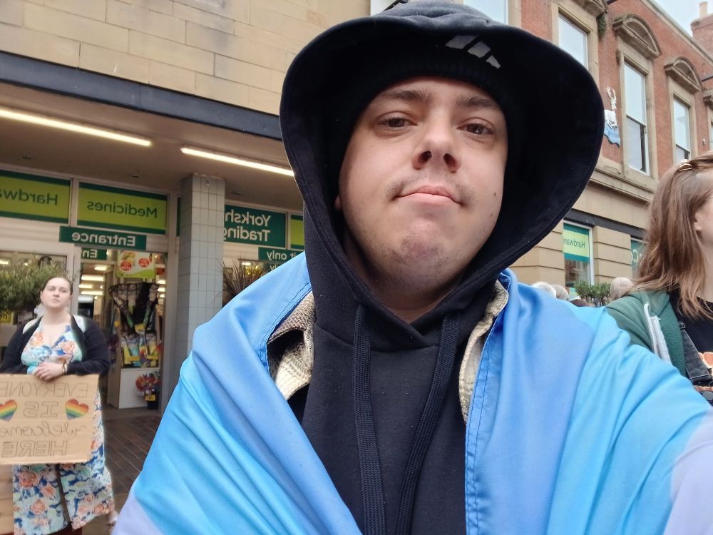 A white transmasc in a black hoodie and beanie with the bigender pride flag around his shoulders 