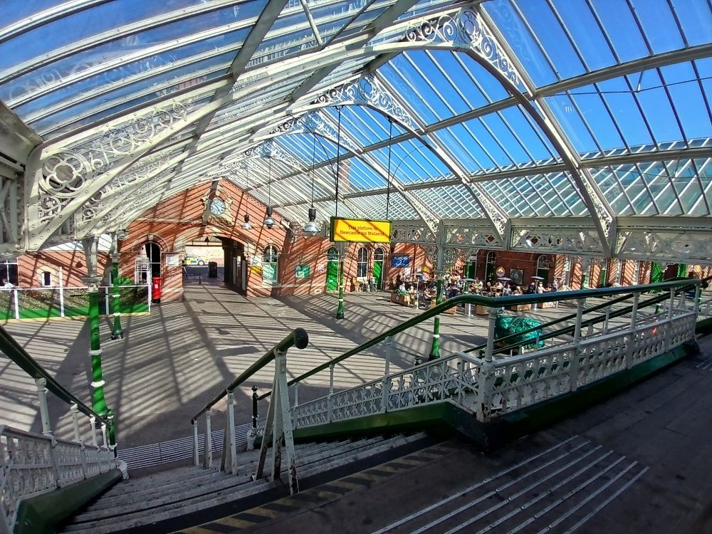 Tynemouth metro station. There is the glass station roof with the blue sky visible thru it, and some people sat at the bar on the station 