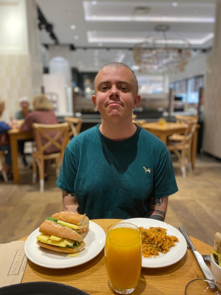 A white transmasc wearing a green t shirt sat at a wooden cafe in a Fenwicks cafe. He has two plates in front of him, one with a cheese sandwich, and one with some rice. He also has some orange juice. He has a shaved head a tattoos on his arms