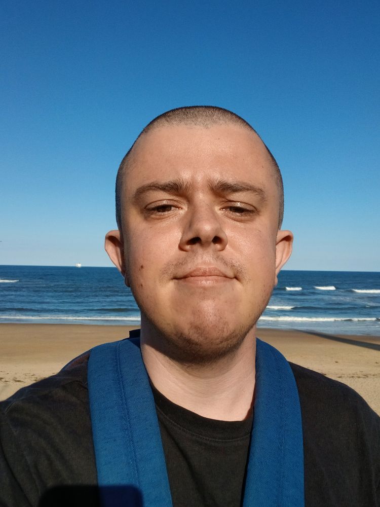 A white transmasc wearing a black t shirt and blue rucksack, and has a shaved head. He's standing on tynemouth longsands beach, with the yellow sand and blue sea visible behind him