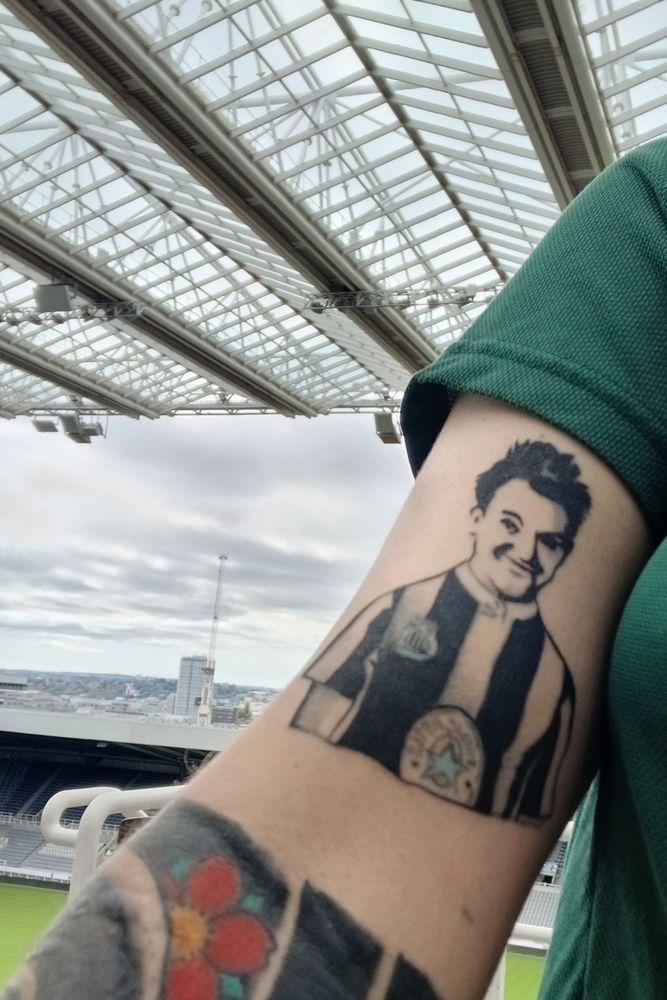 A white arm with a black and white tattoo of Stan Laurel, with the top of a black half sleeve also visible. There is some of a green t shirt visible. In the background is the roof of St James's Park, and the East Stand of St James's, as well as the pitch I'm stood on level 7 of the stadium