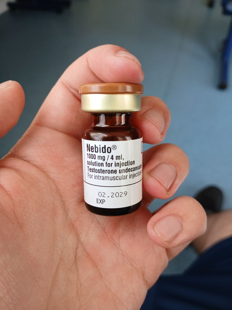 A white hand holding a vial that reads 'nebido 1000mg solution for injection' 