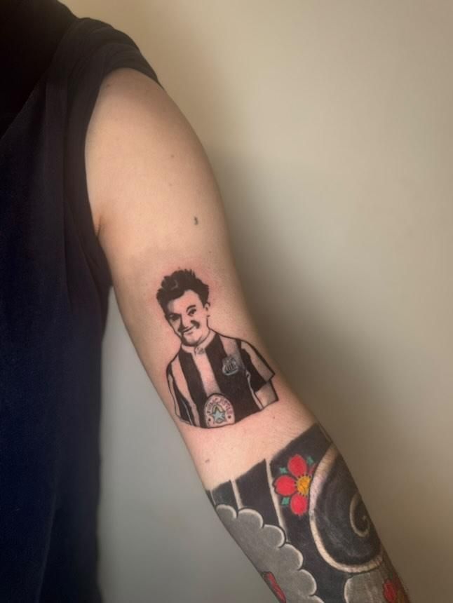 A colour photo of an arm. On the upper arm is a tattoo of stan laurel wearing a vintage Newcastle United shirt. There is a Japanese style half sleeve on the lower arm