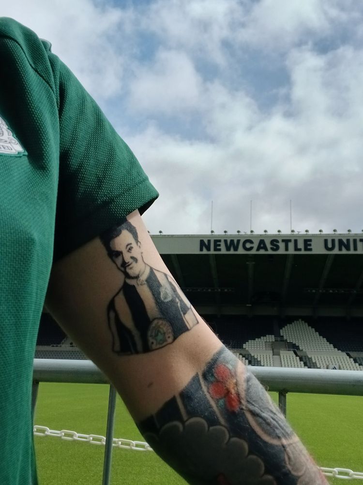The same white arm, tattoos, and green t shirt, but stood in front of the pitch, and East Stand with the 'Newcastle United' signage. There is a metal barrier in front of the pitch