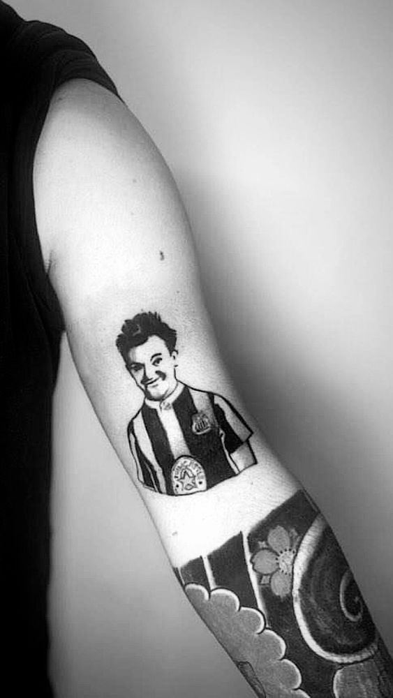 A black and white photo of an arm. On the upper arm is a tattoo of stan laurel wearing a vintage Newcastle United shirt. There is a Japanese style half sleeve on the lower arm