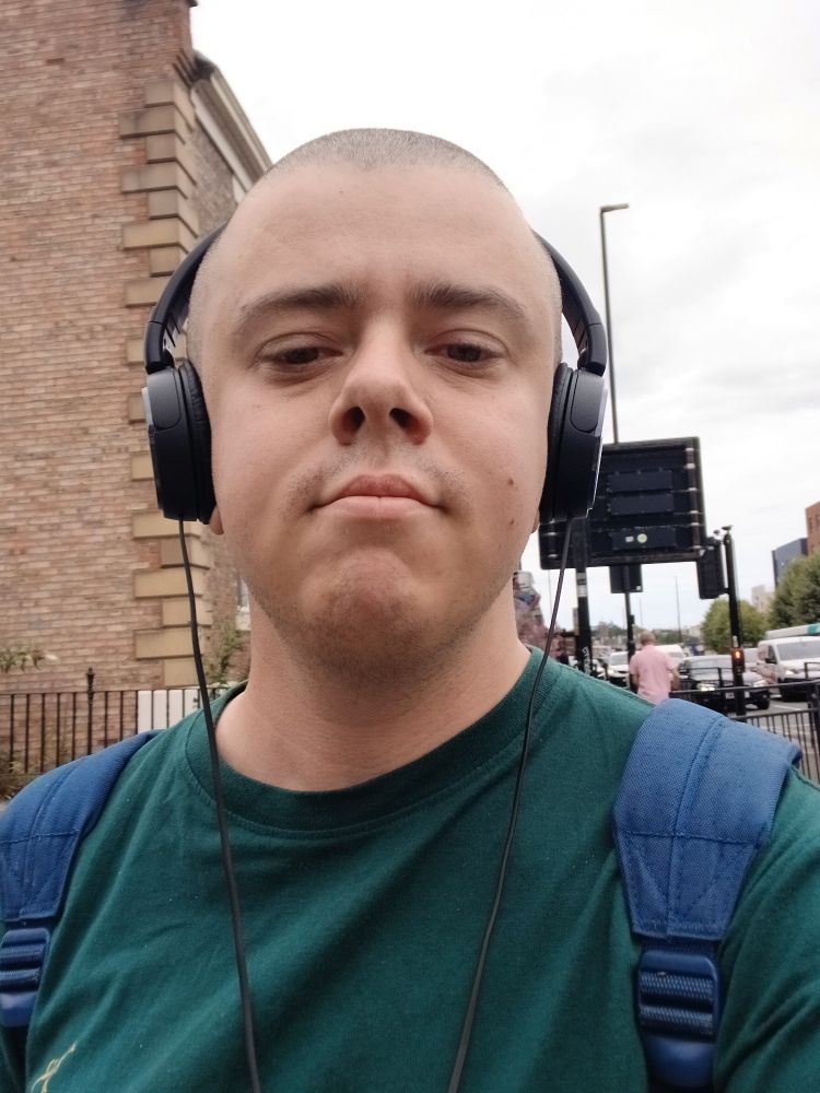 A white transmasc wearing a green t shirt, black headphones, and a blue rucksack