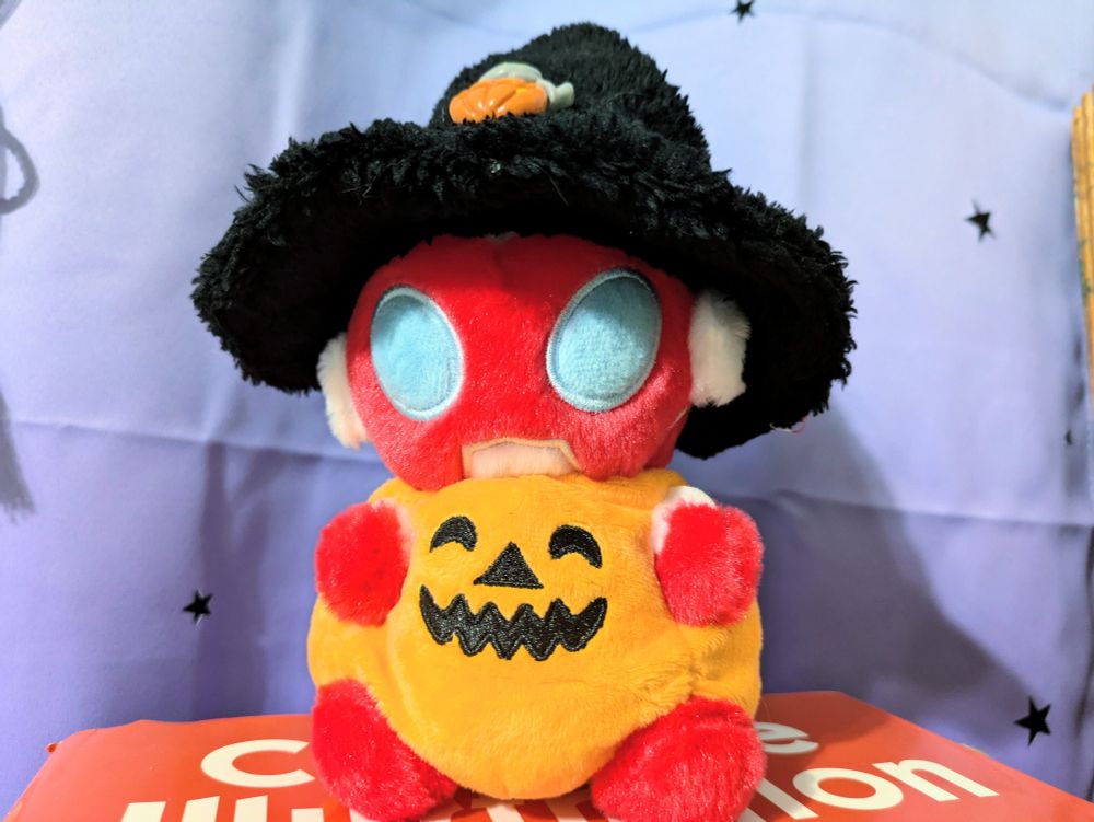 A photo of the Ultraman Omega fuwafuwa doll, dressed in an orange jack-o-lantern halloween costume and fluffy witch hat. End ID.