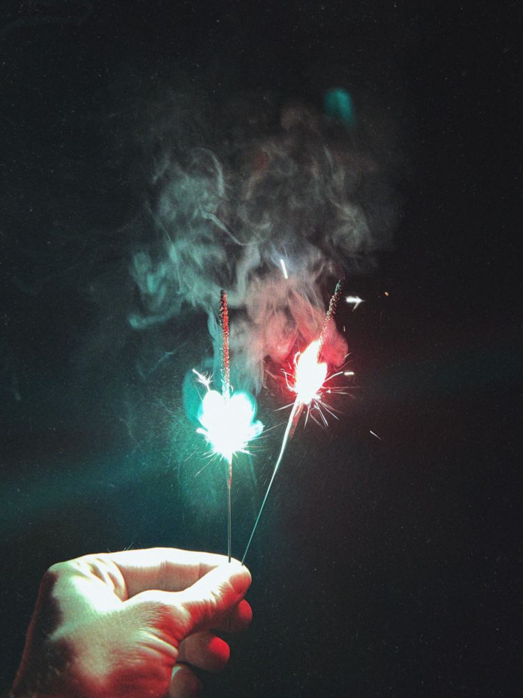 Against a dark background, a hand holds two sparklers — one glowing with a greenish light, the other with a red light, creating bright sparks and swirling smoke