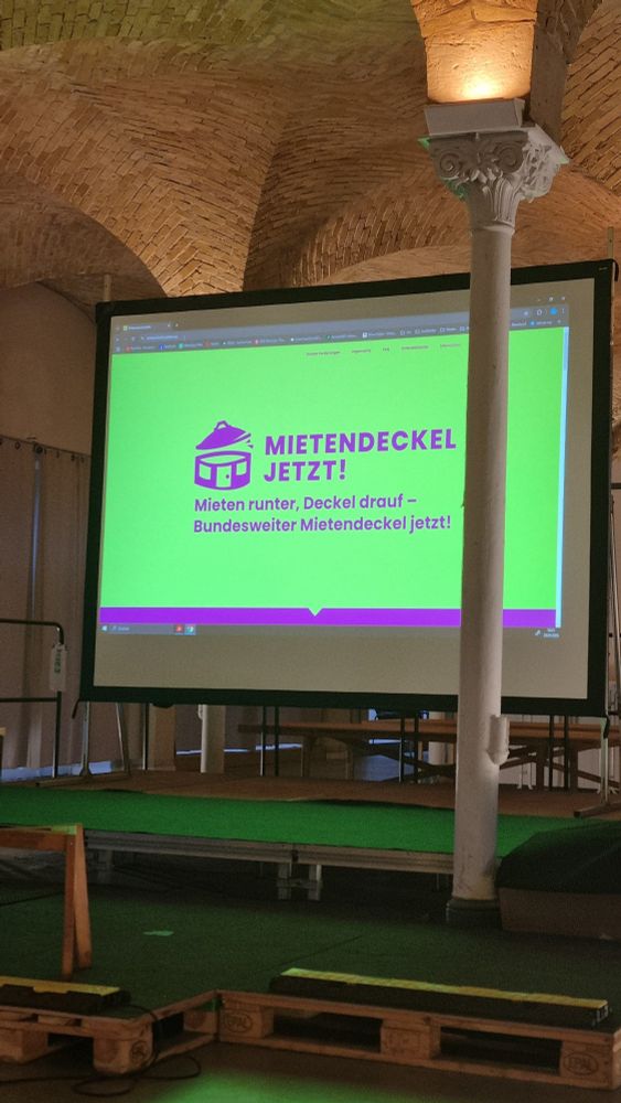 Eine Leinwand, die eine PowerPoint-Folie zeit, mit dem Text: "Mietendeckel jetzt! Mieten runter, Deckel rauf - Bundesweiter Mietendeckel jetzt!"