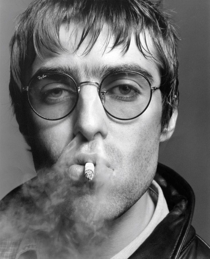 liam gallagher, vocalista da banda oasis. é um homem branco, de cabelo escuro liso curto, olhos esbugalhados claros, sobrancelhas grossas e queixo quadrado. ele está usando óculos transparentes redondos e está fumando um cigarro, expelindo toda a fumaça pelo nariz. foto em preto e branco.