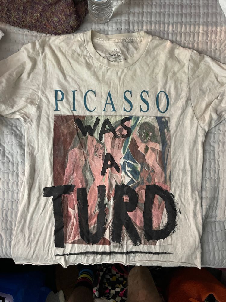 White t-shirt with words “Picasso” on top and a painting by Picasso under it. Drawn on words in black paint say “was a turd” right under Picasso