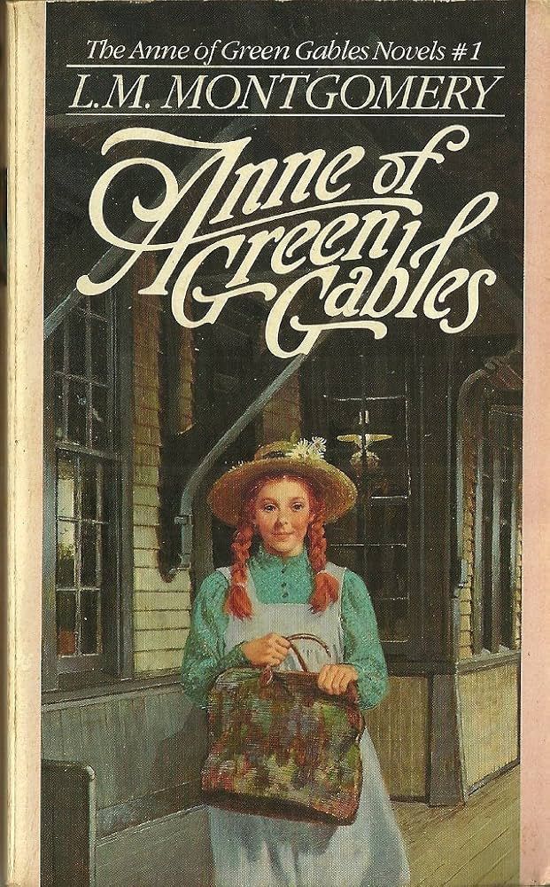 The image is the cover of the book Anne of Green Gables by L.M. Montgomery. It features an illustration of Anne Shirley, a young girl with red hair in braids, wearing a straw hat adorned with flowers, a green blouse, and a light blue pinafore. She holds a patterned carpet bag in her hands. The setting appears to be outside a rustic building with wooden walls and large windows. The title, Anne of Green Gables, is written in large, decorative white script, with "The Anne of Green Gables Novels #1" and the author's name, "L.M. Montgomery," in smaller text above.